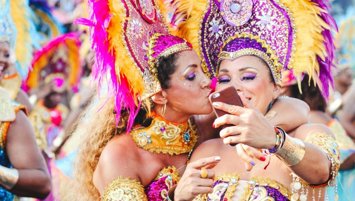 Two women Carnival dancers pose for a selfie as one kisses other on the cheek.