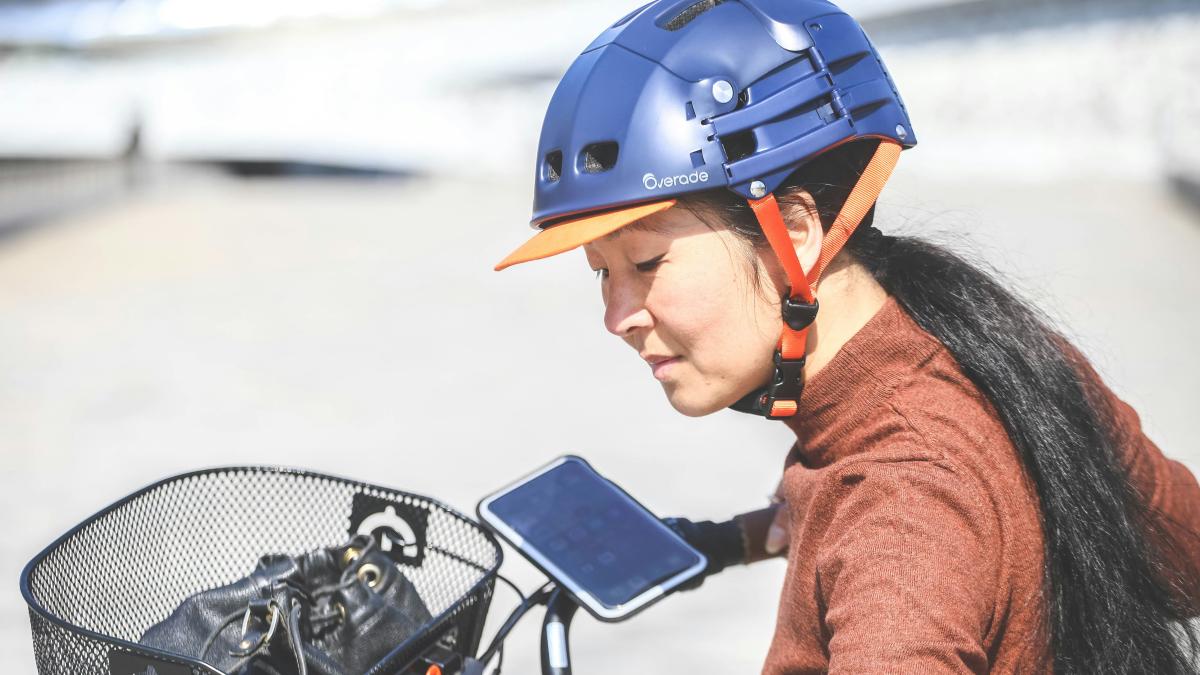 Jeune femme asiatique sur son vélo électrique. Elle porte un casque pliable bleu et une visière orange. Son téléphone est accroché à son guidon sur un support magnétique.