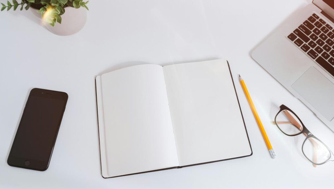 An open empty notebook on a white desk next to an iPhone and a MacBook