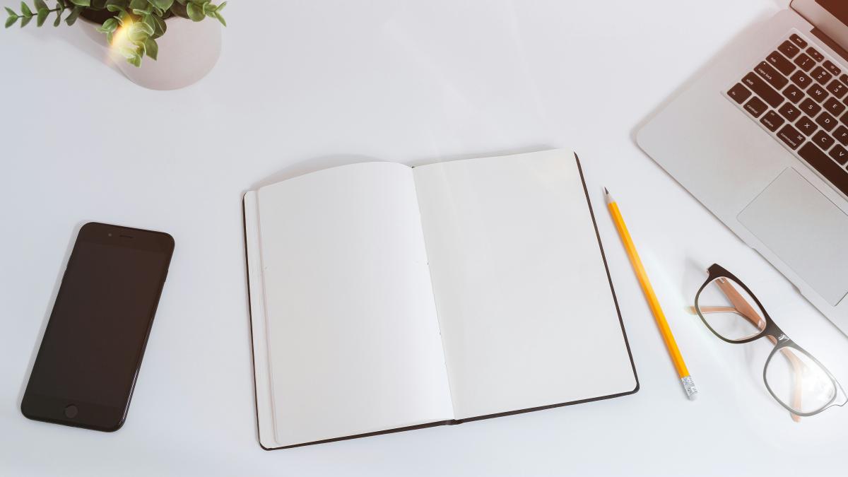 An open empty notebook on a white desk next to an iPhone and a MacBook