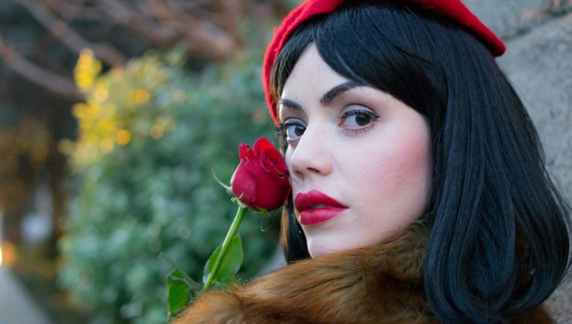 woman wearing brown coat holding red rose near wall during daytime