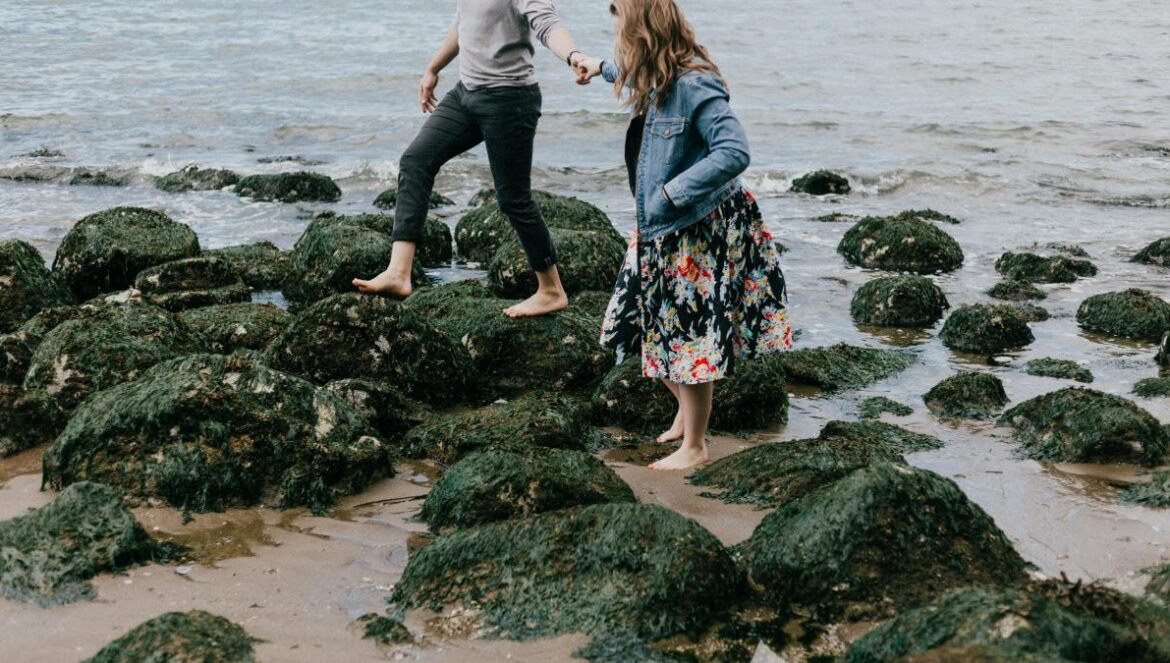 man and woman holding hand together while stepping on rocks near sea