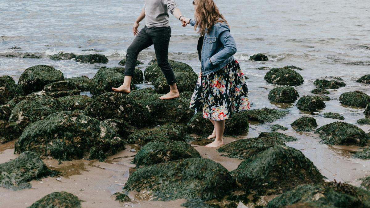 man and woman holding hand together while stepping on rocks near sea
