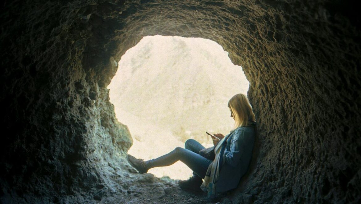 Woman sits in a cave, looking at a phone.
