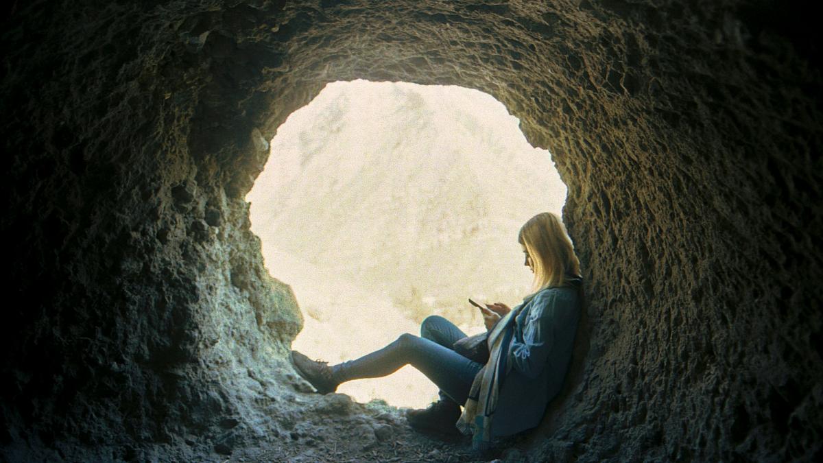 Woman sits in a cave, looking at a phone.