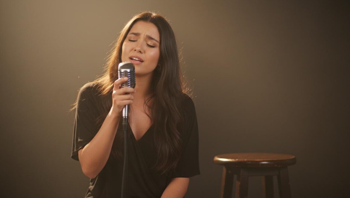 Joven cantante sobre un pequeño escenario, cantando al micrófono bajo una luz cálida, con fondo desenfocado.